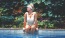 woman in bathing suit and sunglasses sits on pool ledge with her legs in the water